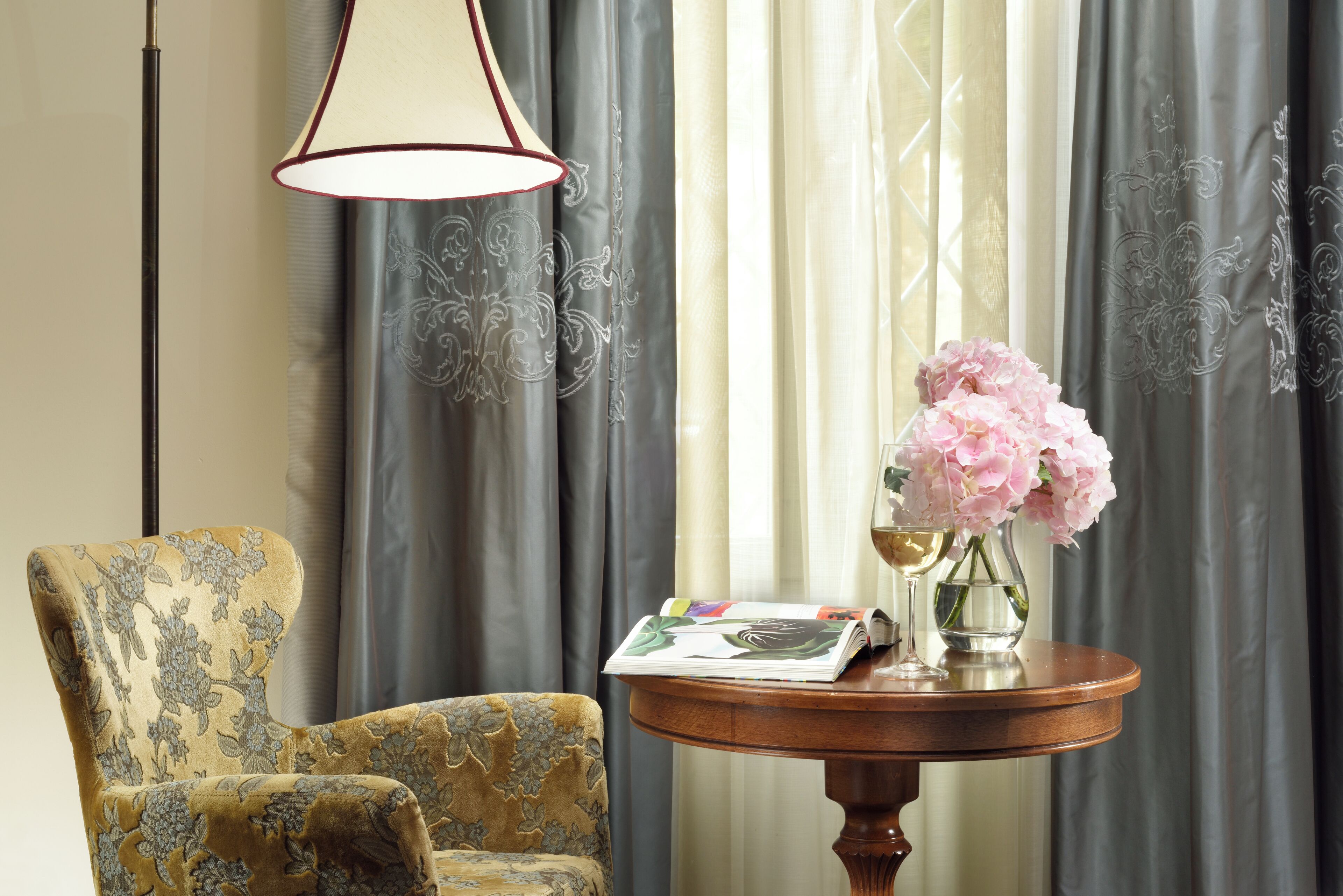 Una habitación con un sillón estampado, una pequeña mesa redonda de madera con una revista y un jarrón de flores, y una lámpara.