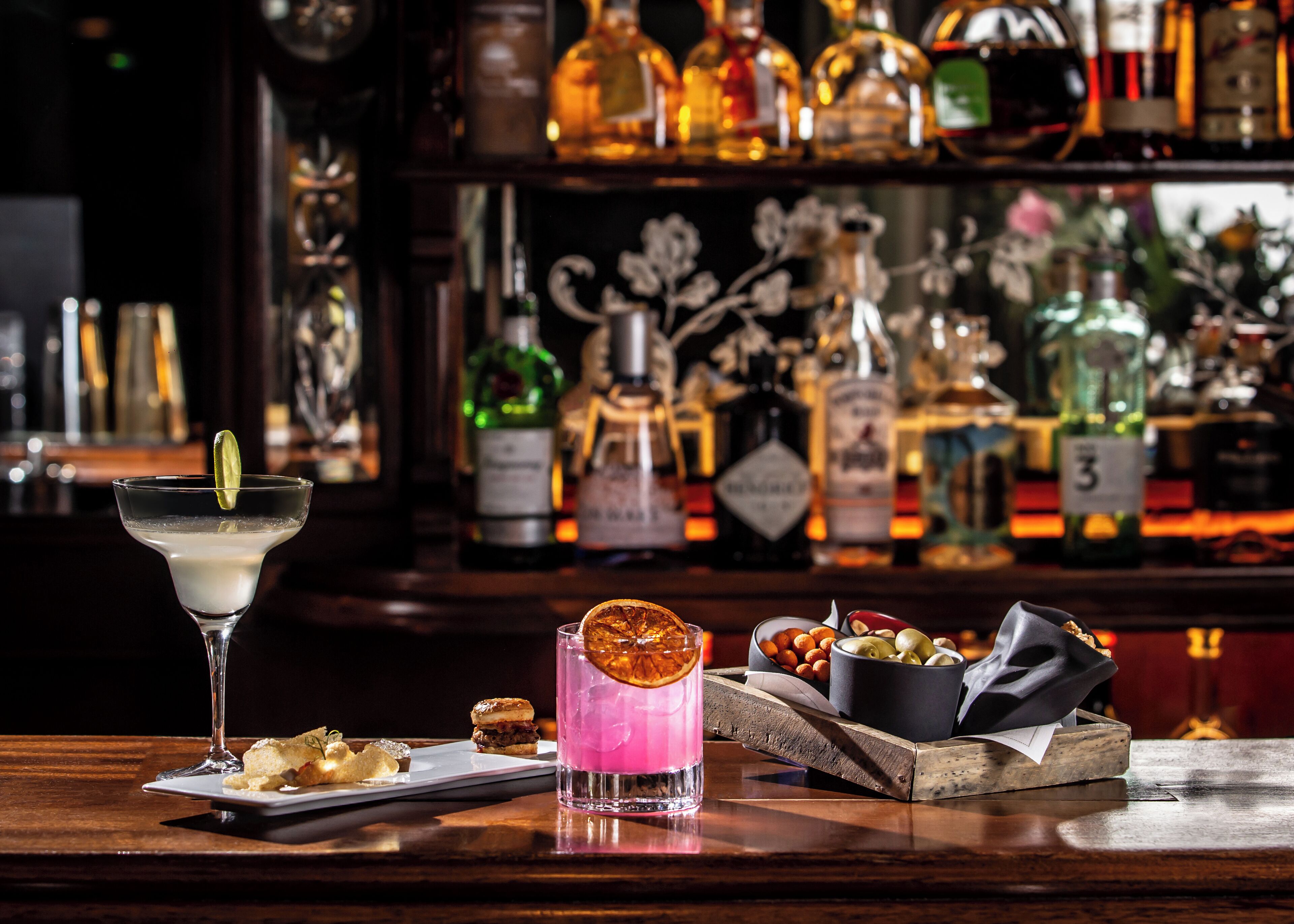 Een barsetting met een martini-glas, een roze cocktail en een schaal met noten en gedroogd fruit.