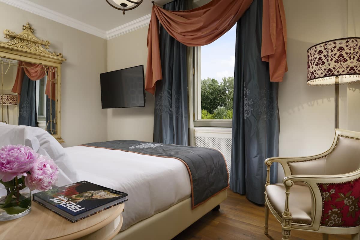 A bedroom with a bed, a chair, a TV, a mirror, and a window with curtains.