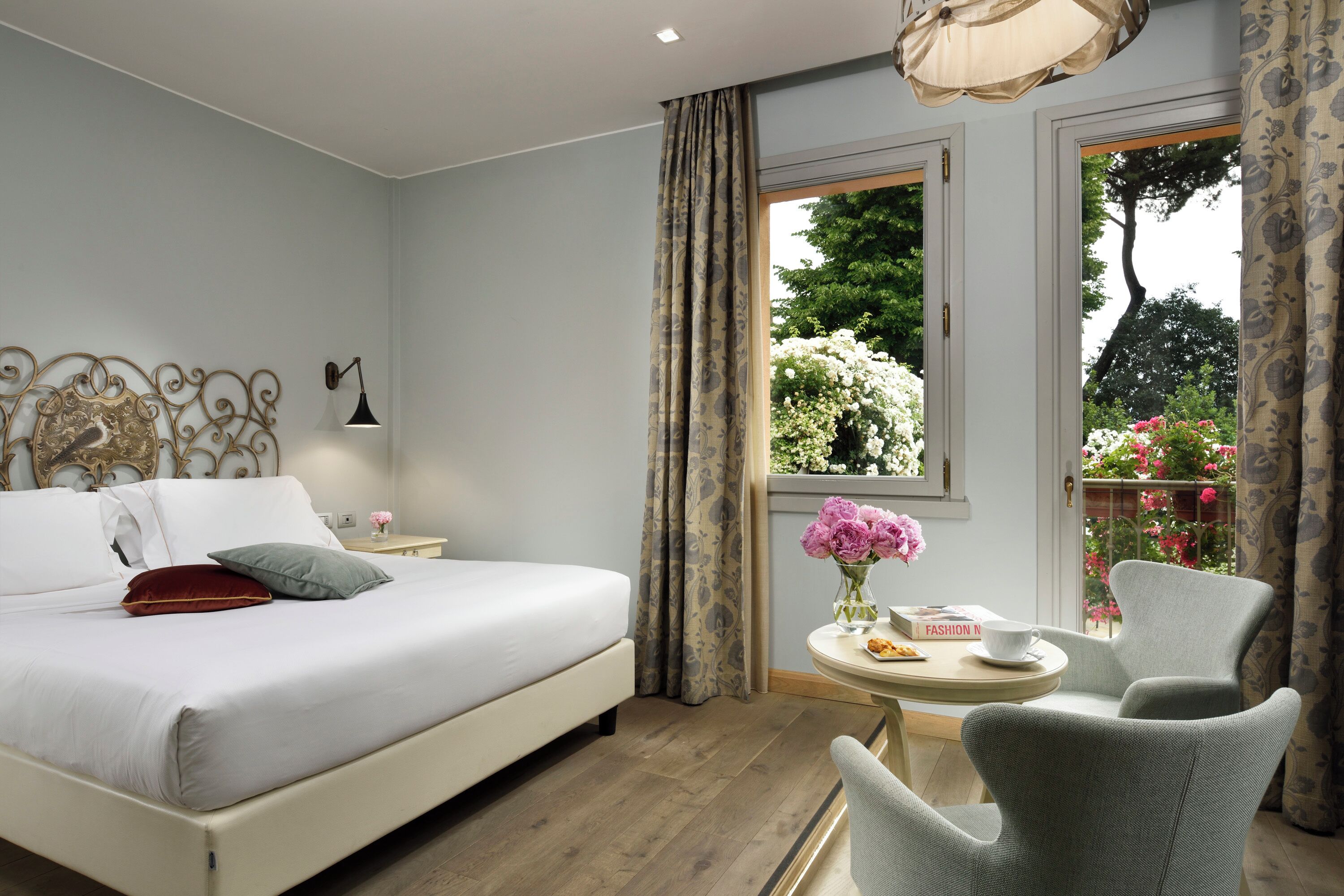 A bedroom with a bed, a bedside table with a lamp, a window with curtains, a small round table with a vase of flowers, and a chair.