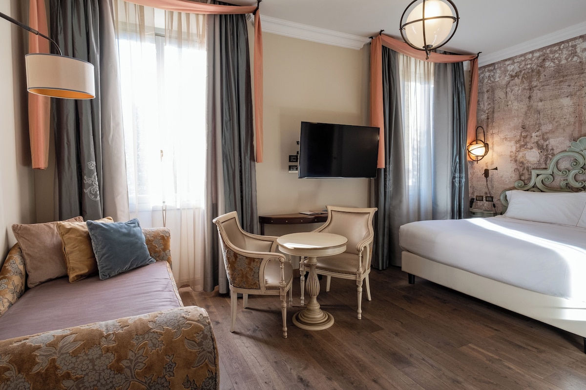 A hotel room with a bed, a sofa, a dining table with chairs, a TV, and a lamp.