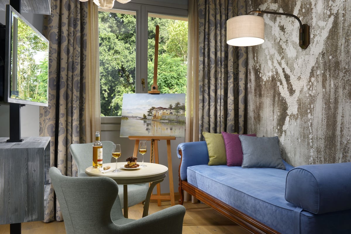 A room with a blue sofa, a small round table with a bottle and glasses, a painting on the wall, and a window with curtains.