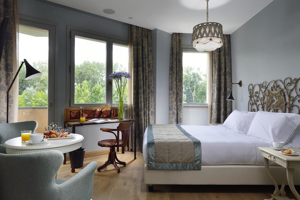 A hotel room with a bed, a desk with breakfast, a chair, a lamp, and large windows.