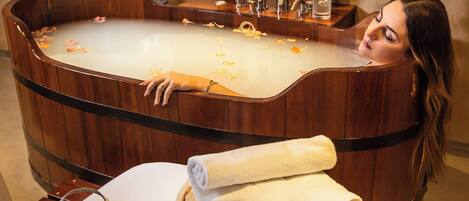 A woman relaxing in a wooden bathtub with candles, surrounded by towels and a tray of flowers.