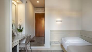Un dormitorio con cama, mesita de noche, silla y escritorio con otra silla. Hay una puerta que da a otra habitación y un equipo de aire acondicionado en la pared.