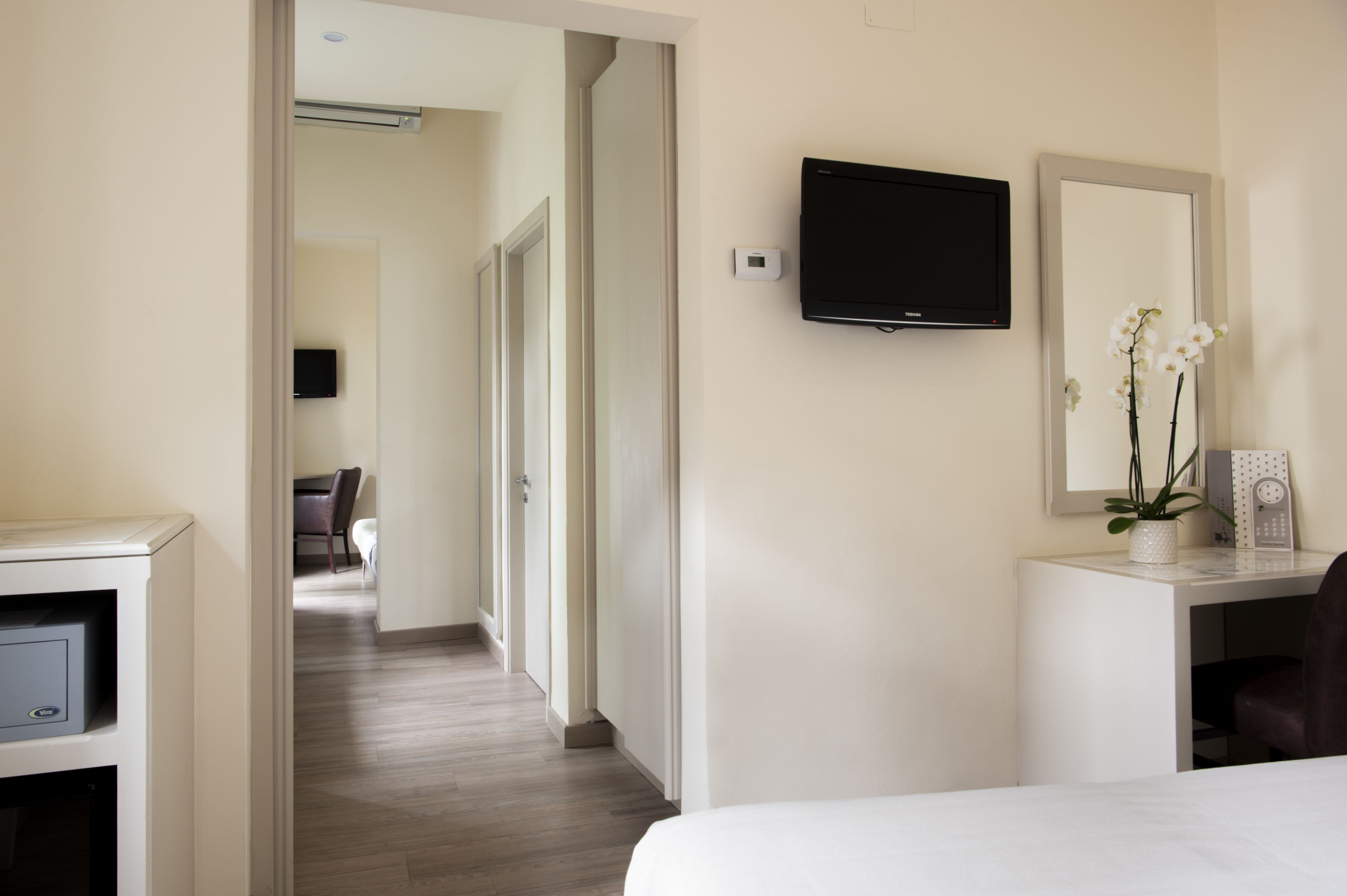 A hotel room with a bed, a desk with a flat-screen TV, a mirror, and a potted orchid.
