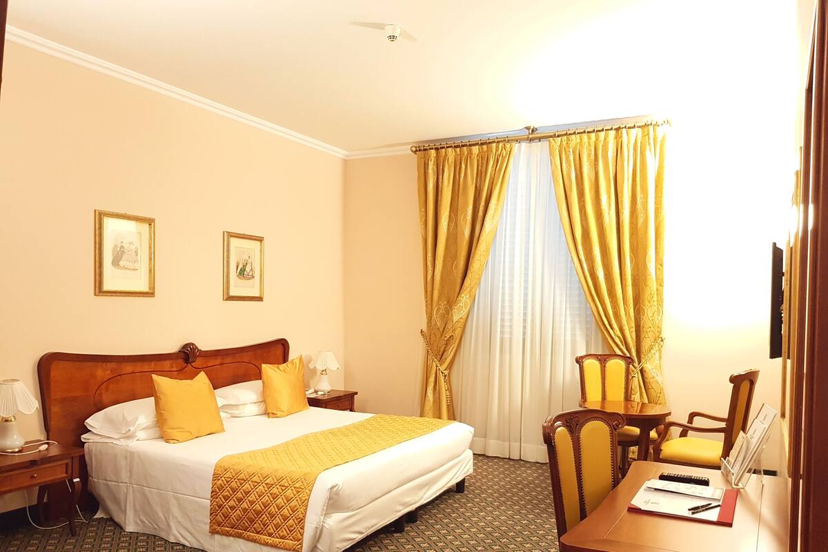 Ein Hotelzimmer mit einem großen Bett, zwei Nachttischen, einem Schreibtisch mit Stuhl und einem Fenster mit Vorhängen.