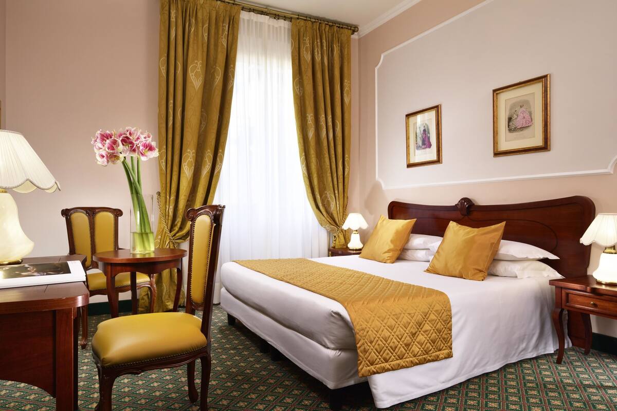 Ein Hotelzimmer mit einem großen Bett, einem Schreibtisch, einem Stuhl, einer Vase mit Blumen und zwei gerahmten Bildern an der Wand.