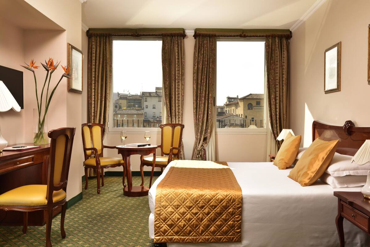 Ein Hotelzimmer mit einem Bett, zwei Sesseln, einem Schreibtisch, einer Lampe und einem Fenster mit Vorhängen.