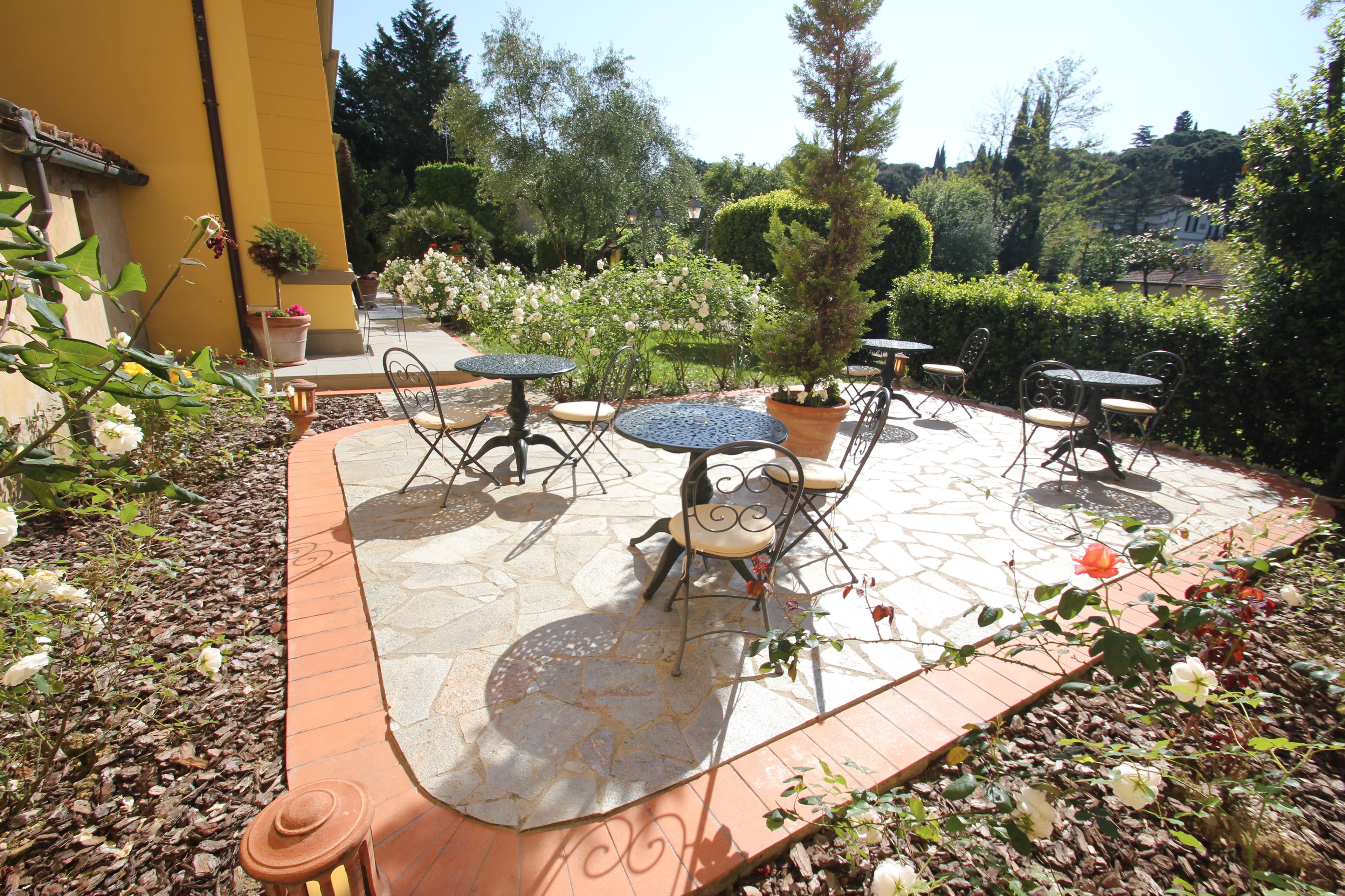 An outdoor patio with stone flooring, metal tables and chairs, surrounded by potted plants and flowers.