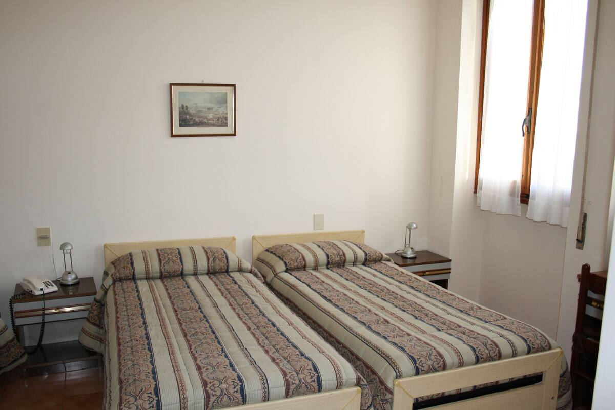 Una habitación con dos camas, una mesita de noche con lámpara, un cuadro enmarcado en la pared y una ventana con cortinas.