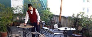 A person serving at an outdoor café with multiple tables and chairs, surrounded by greenery.