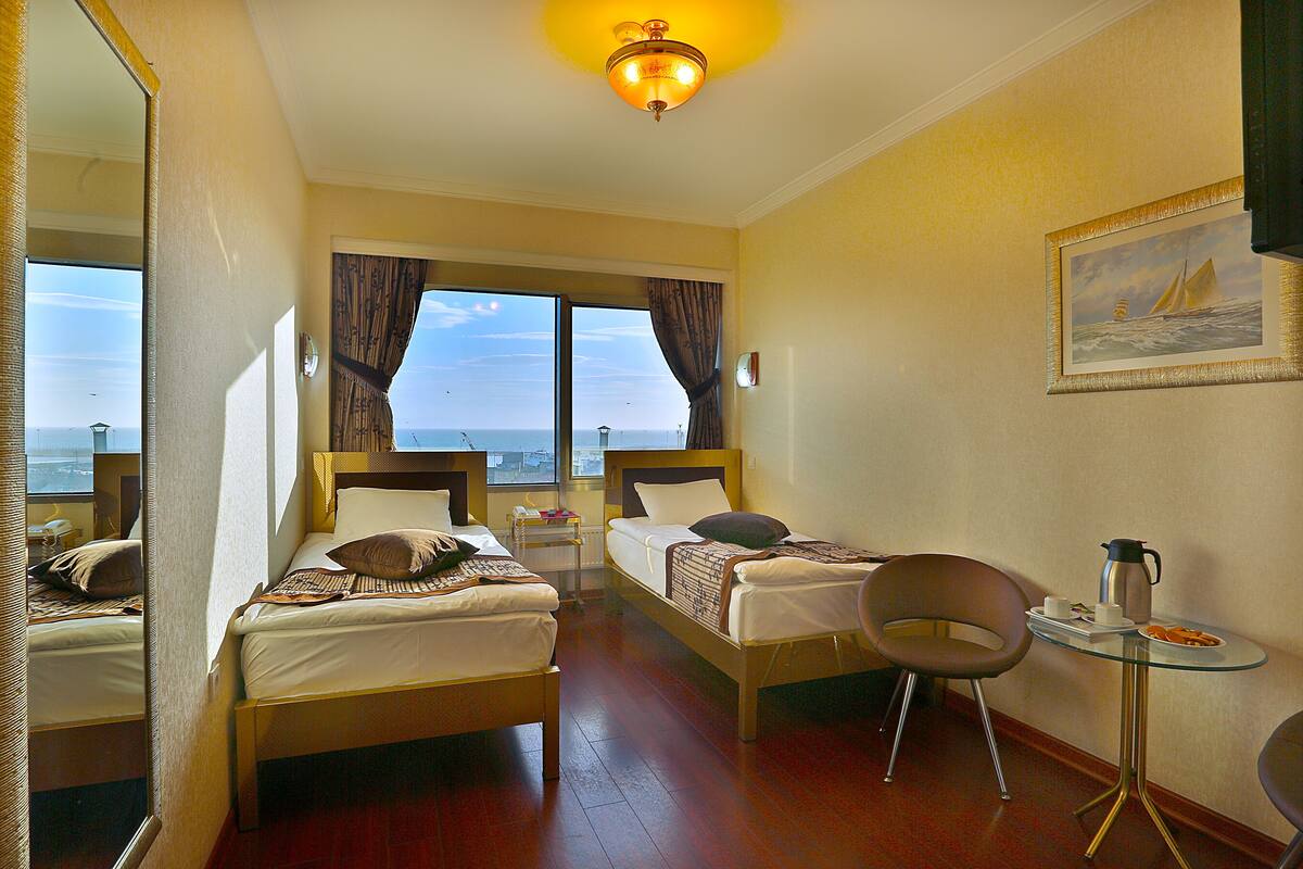 A hotel room with two beds, a small table with a chair, a painting on the wall, and a view of the sea through the window.