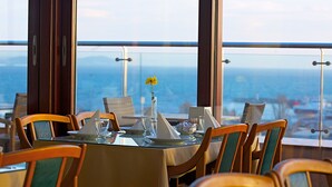 Une table à manger dressée pour quatre personnes, avec des chaises, un vase contenant une fleur jaune et une vue sur la mer par de grandes fenêtres.