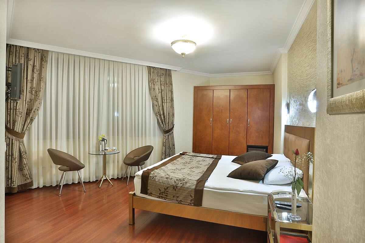 A hotel room with a bed, two chairs, a small table, a wardrobe, and a large window with curtains.