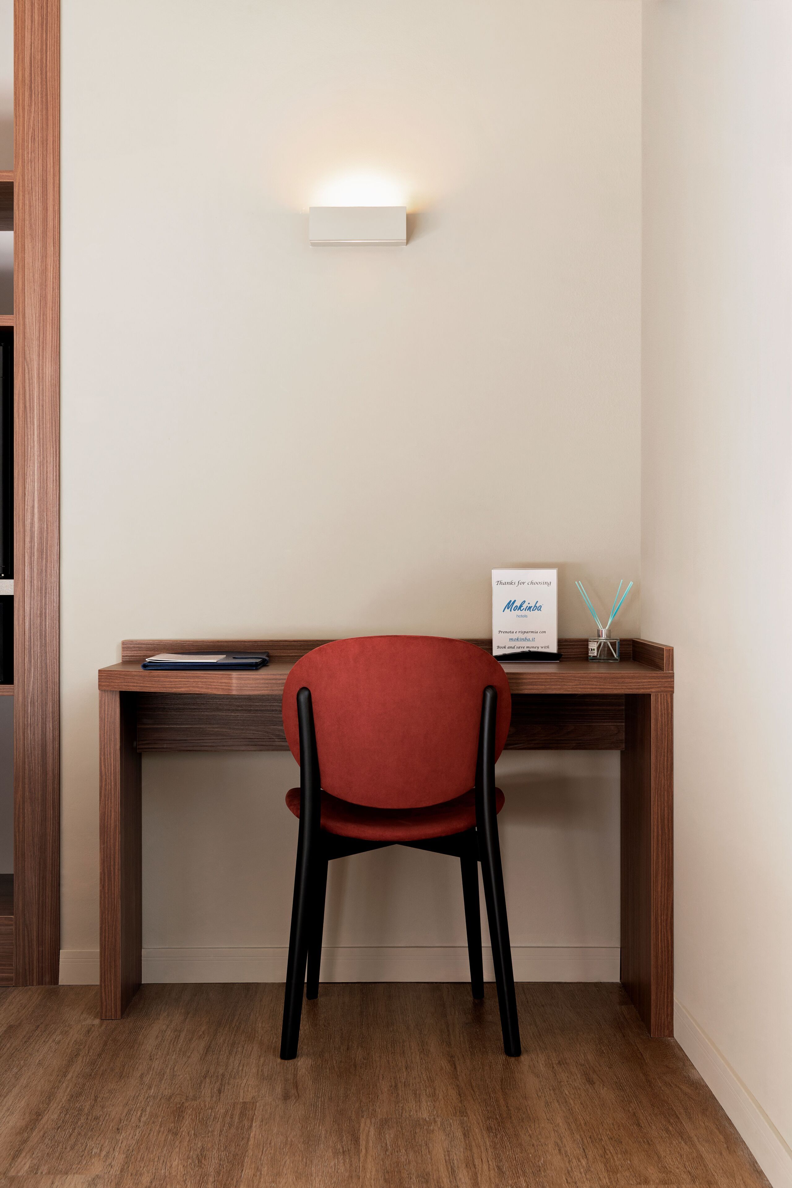 Un bureau en bois avec un ordinateur portable et une pancarte, une chaise rouge et un luminaire fixé au mur.