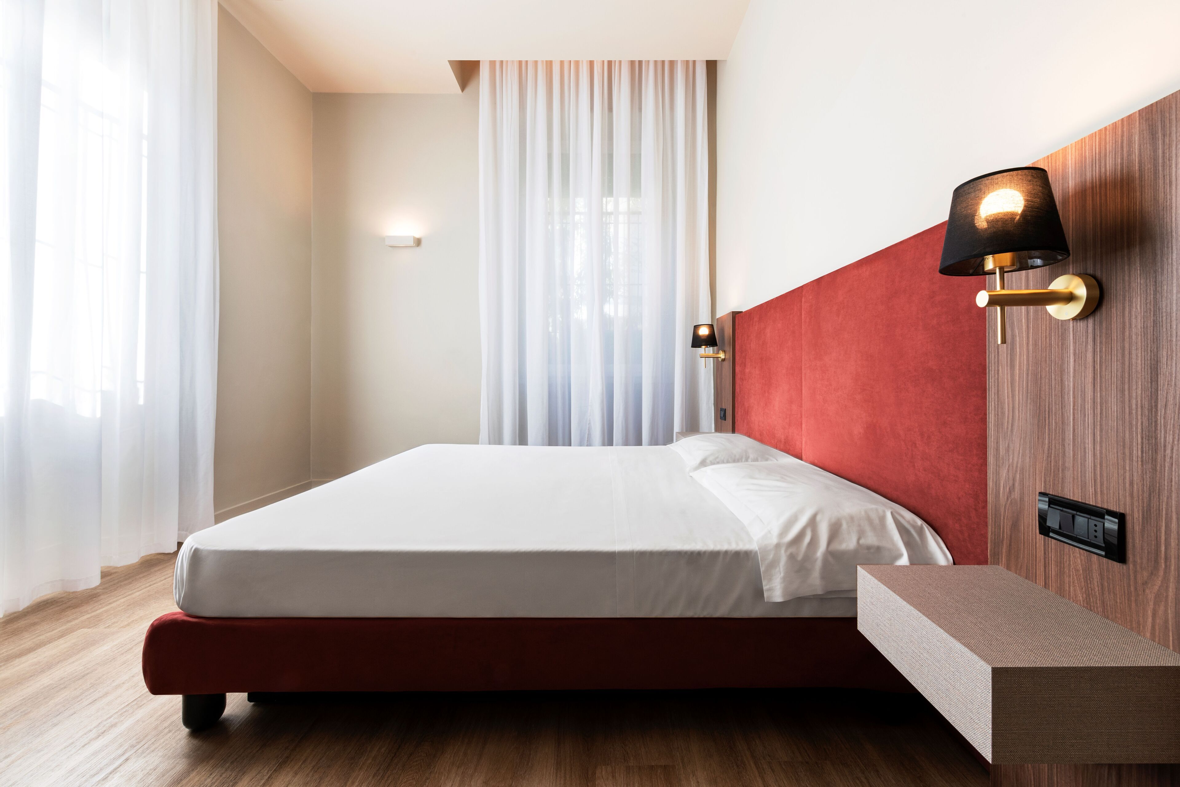 A neatly made bed with a red headboard, a bedside table with a lamp, and a window with sheer curtains.