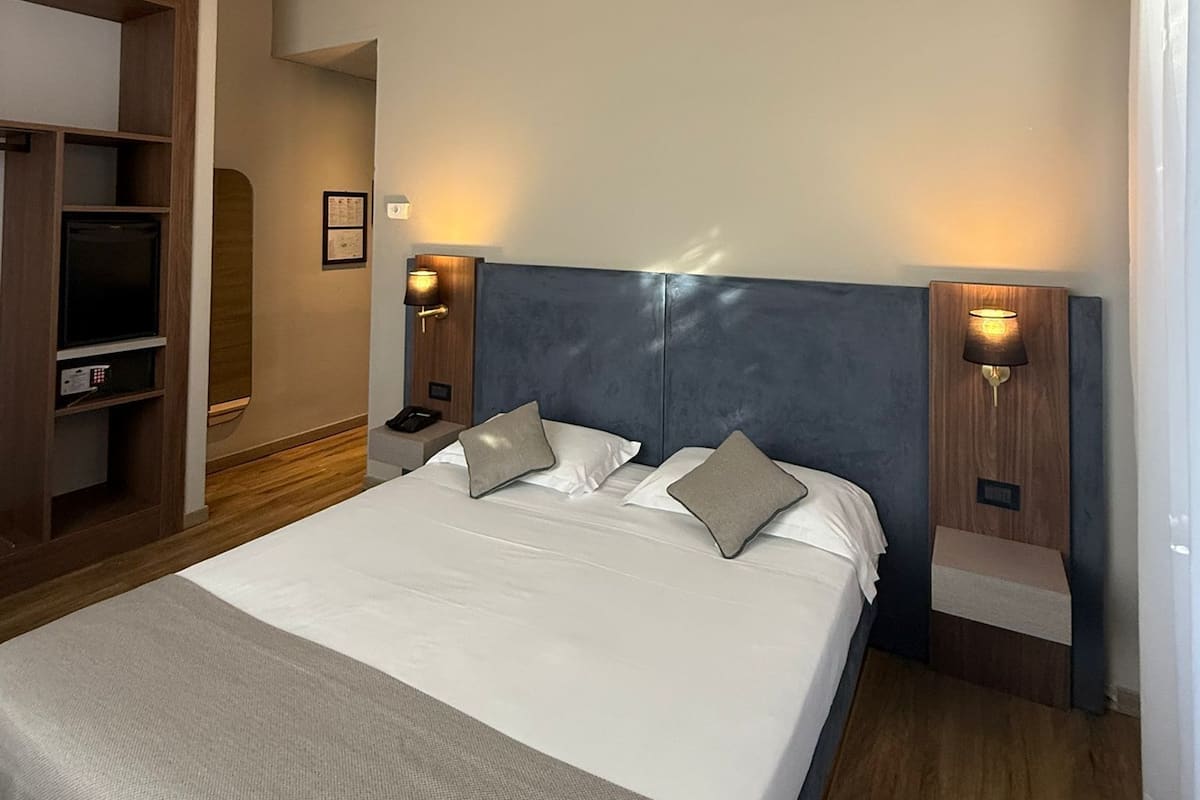 A modern hotel room with a large bed, wooden headboard, two bedside tables with lamps, a built-in TV, and a wardrobe.