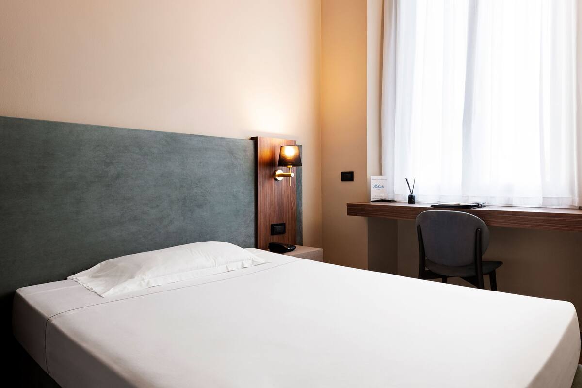A hotel room with a bed, a desk, a chair, a lamp, and a window with curtains.