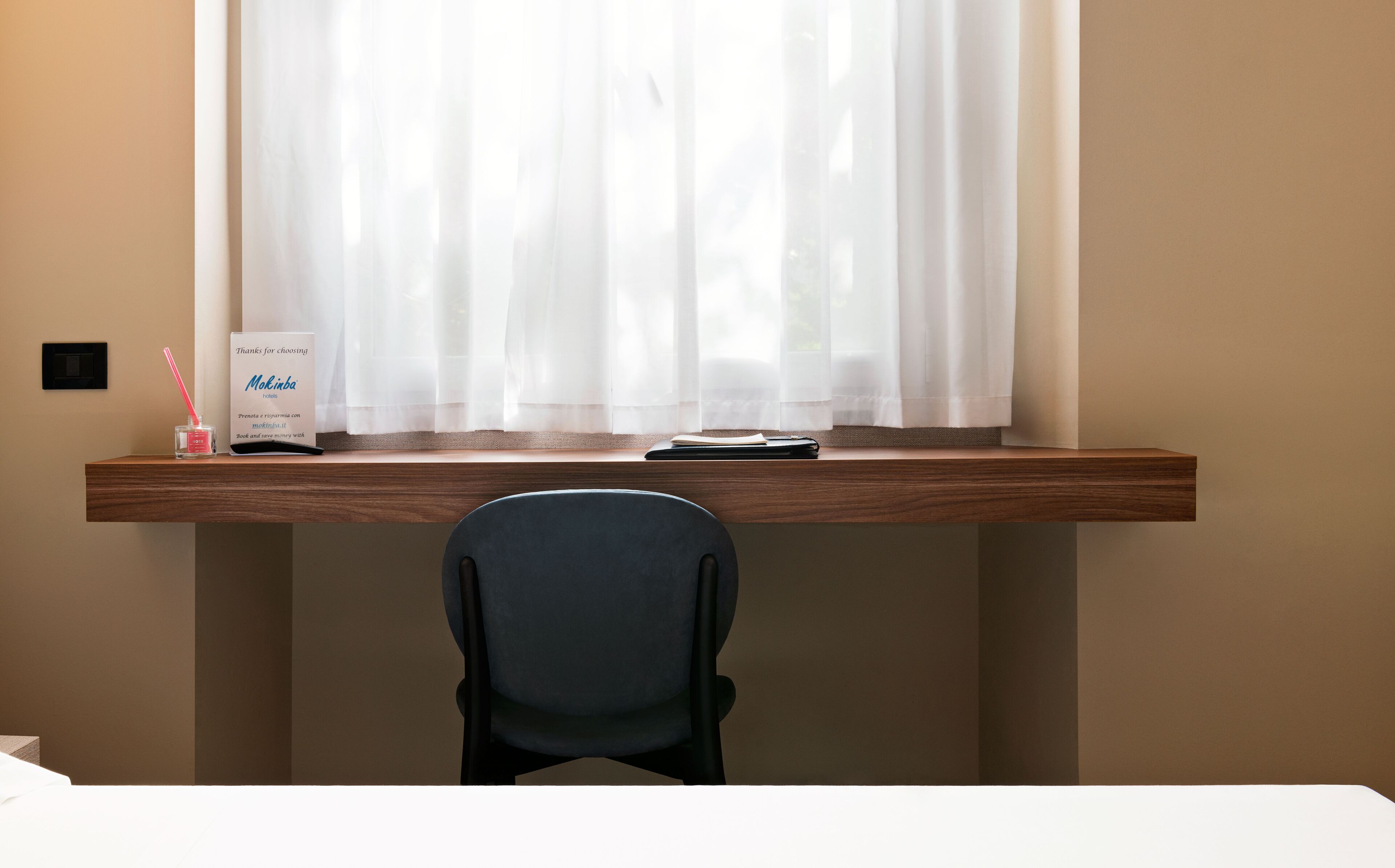 A hotel desk with a chair, a sign, and a window with sheer curtains.