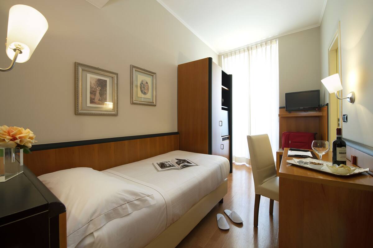 A hotel room with a bed, a desk with a laptop, a chair, a TV, and a wine bottle.