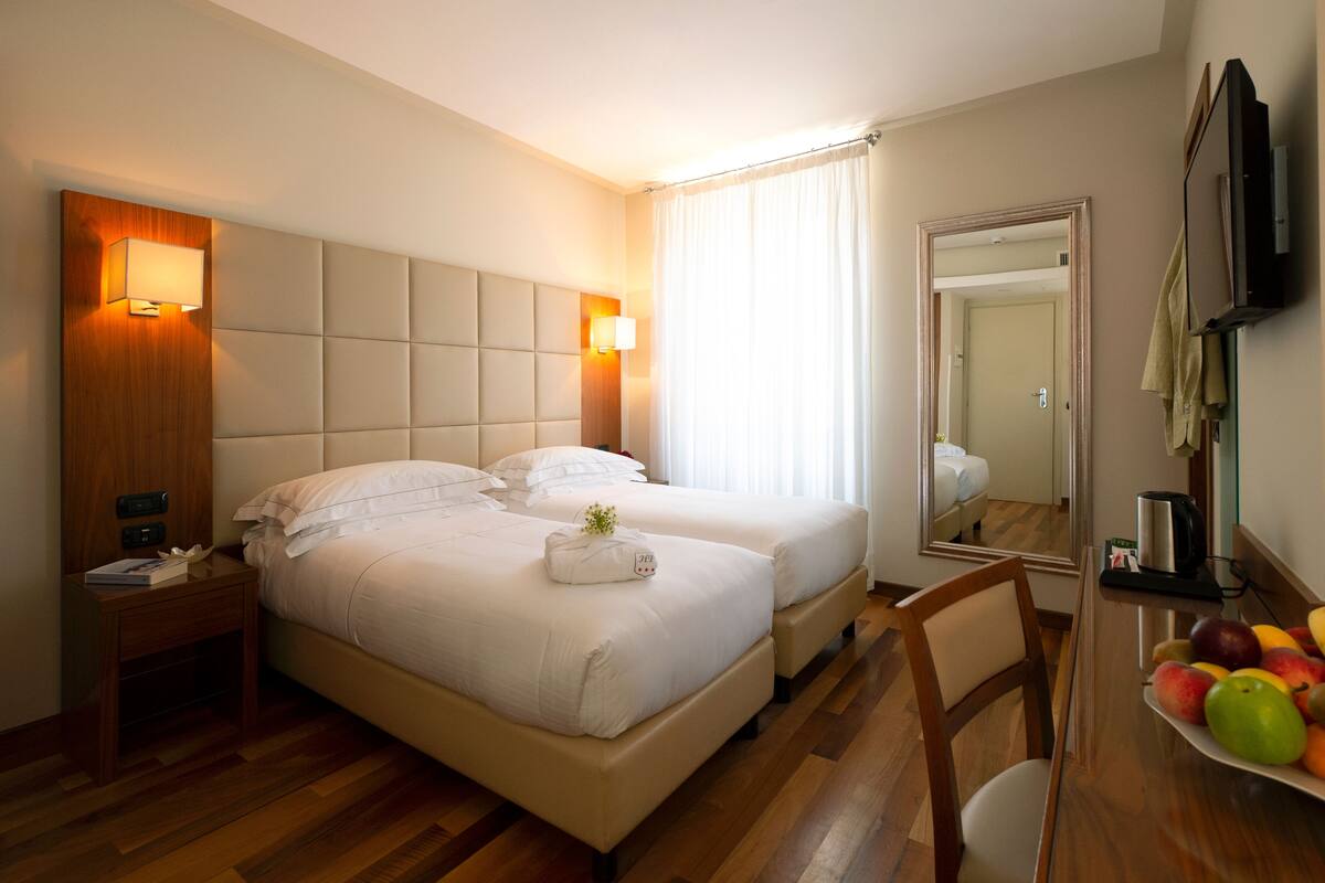 A hotel room with two beds, a wooden headboard, bedside lamps, a mirror, a wooden floor, and a fruit bowl on a table.