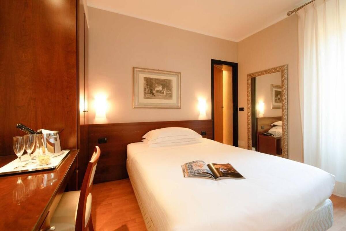 A hotel room with a neatly made bed, a bedside table with glasses and a bottle, a framed picture on the wall, and a large mirror.