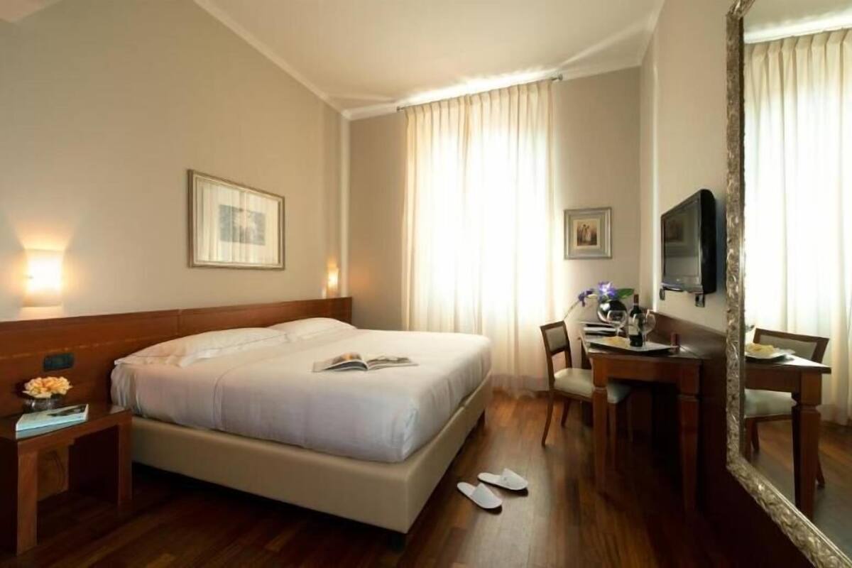 A hotel room with a bed, bedside table, lamp, desk, chair, TV, and curtains.