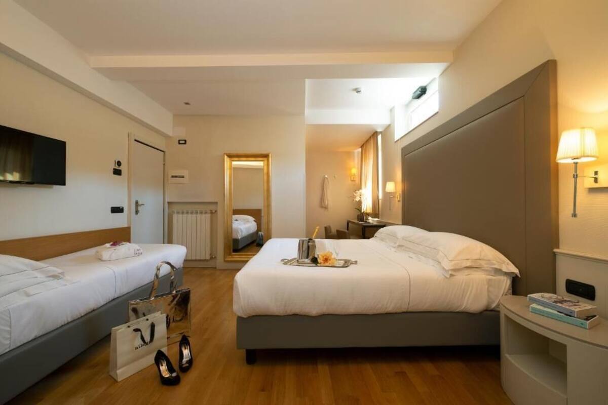 A hotel room with two beds, a TV, a bedside table with a lamp, and a view of another room through a door.