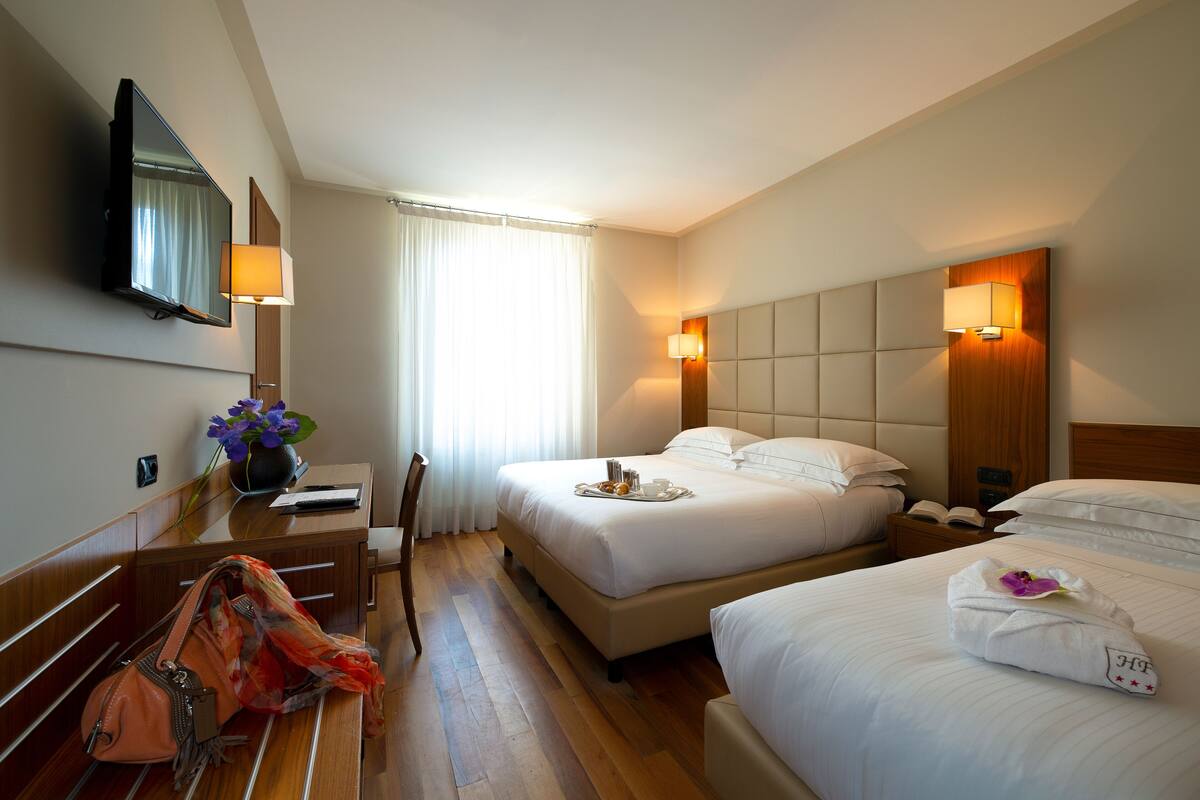 A hotel room with two beds, a desk with a lamp, a TV, and a window with curtains.