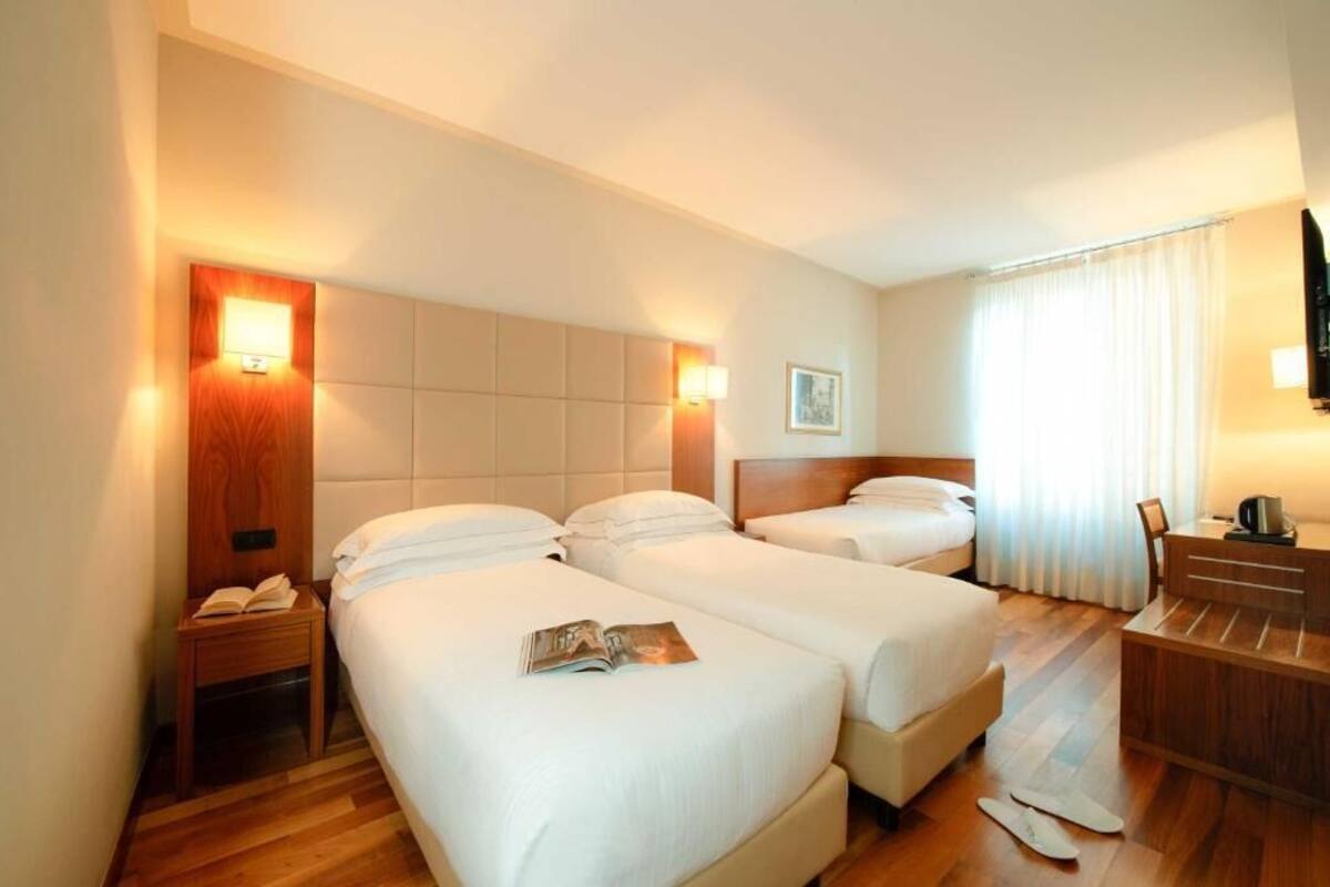 A hotel room with two beds, a wooden headboard, bedside tables with lamps, a desk with a chair, and a window with curtains.