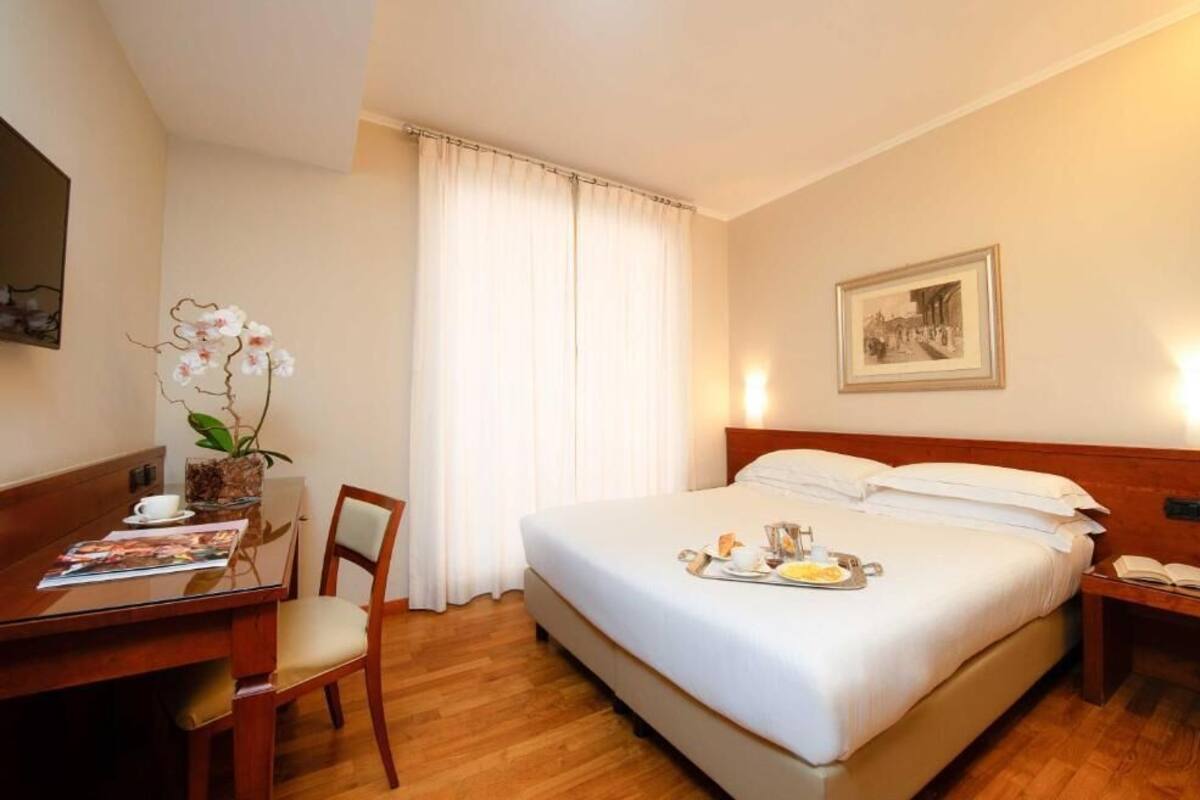 A hotel room with a bed, a desk with a chair, a dining table with a tray of breakfast, a potted orchid, and a framed picture on the wall.