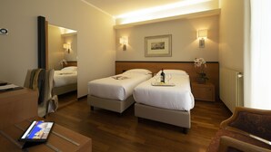 Habitación de hotel con dos camas, un escritorio con una computadora portátil, una silla y una mesita con una bandeja de vino y copas.