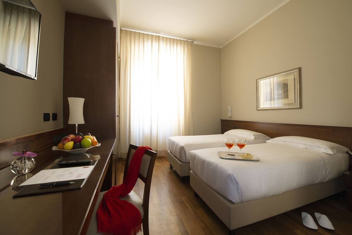 A hotel room with two beds, a desk with a bowl of fruit, a remote control, and a tray with glasses and a plate.