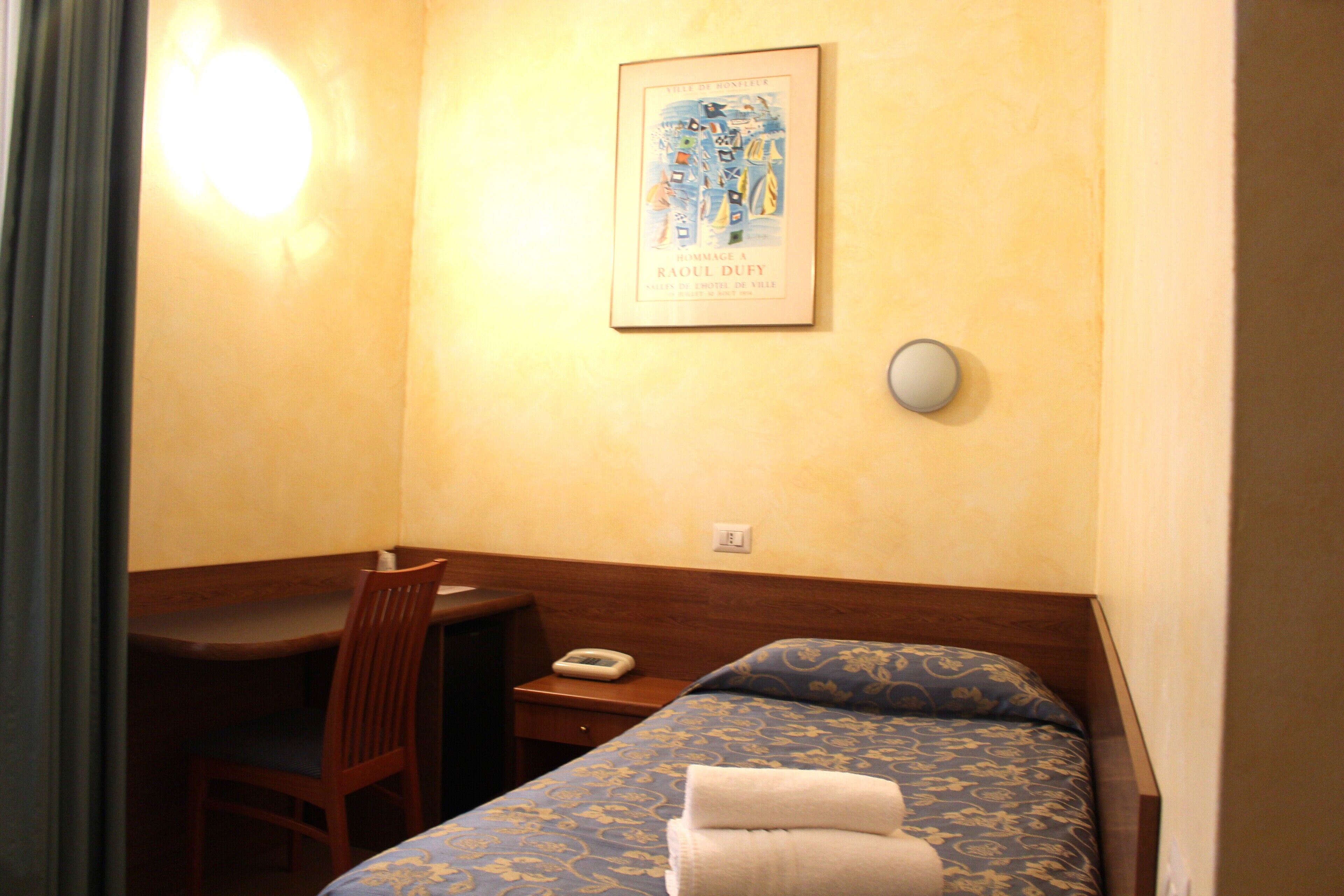 Une chambre d’hôtel comprenant un lit, un bureau, une chaise, un tableau encadré au mur et une table de chevet avec une lampe.