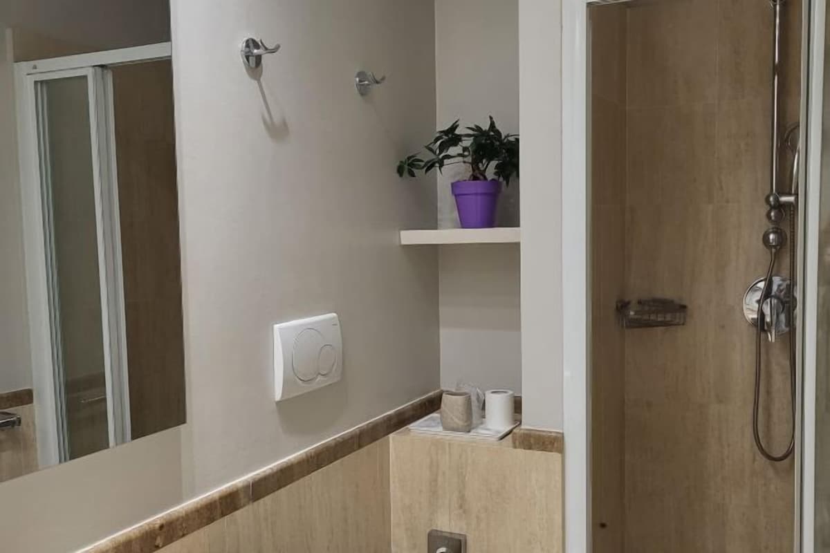 Un baño con ducha, inodoro y lavabo. Hay una planta en maceta sobre un estante encima del lavabo.