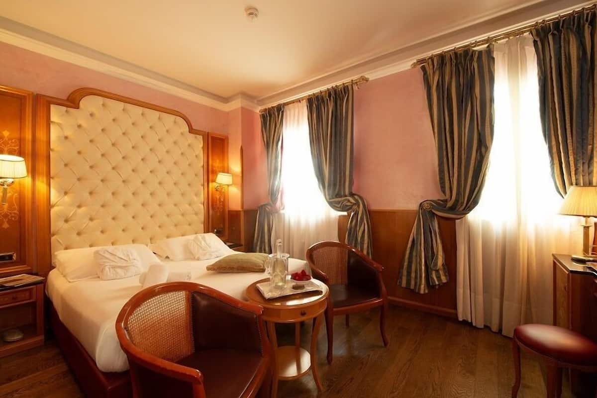 A hotel room with a large tufted headboard, a bed with white linens, two wooden chairs, a small round table with a tray, and curtains on the windows.