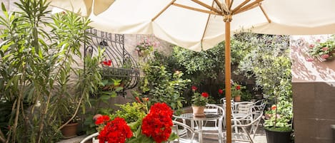 An outdoor patio area with a table and chairs under a canopy, surrounded by potted plants and a brick wall.