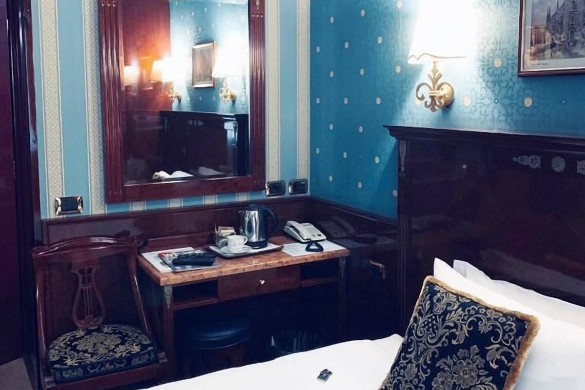 A hotel room with a bed, a desk with a telephone, a kettle, and a chair.