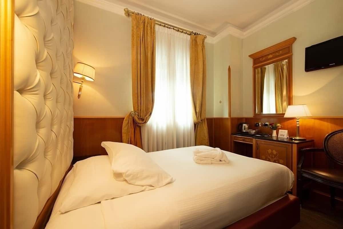 A hotel room with a large bed, a wooden headboard, a bedside table with a lamp, a mirror, and a television on the wall.