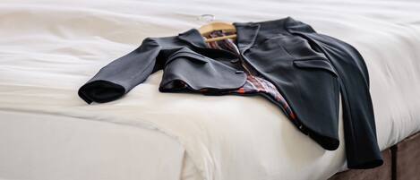 A neatly made bed with a black jacket on top, a wooden headboard, a wall-mounted lamp, and a window with curtains.