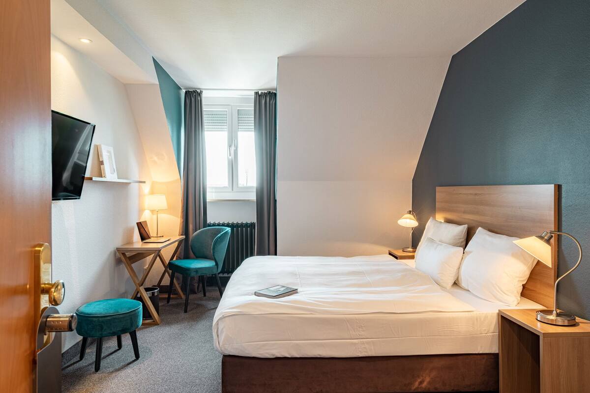 A hotel room with a bed, bedside table, lamp, chair, desk, TV, and a window with curtains.