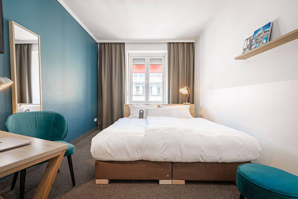 A hotel room with a bed, a desk, a chair, a bedside table with a lamp, and a window with curtains.