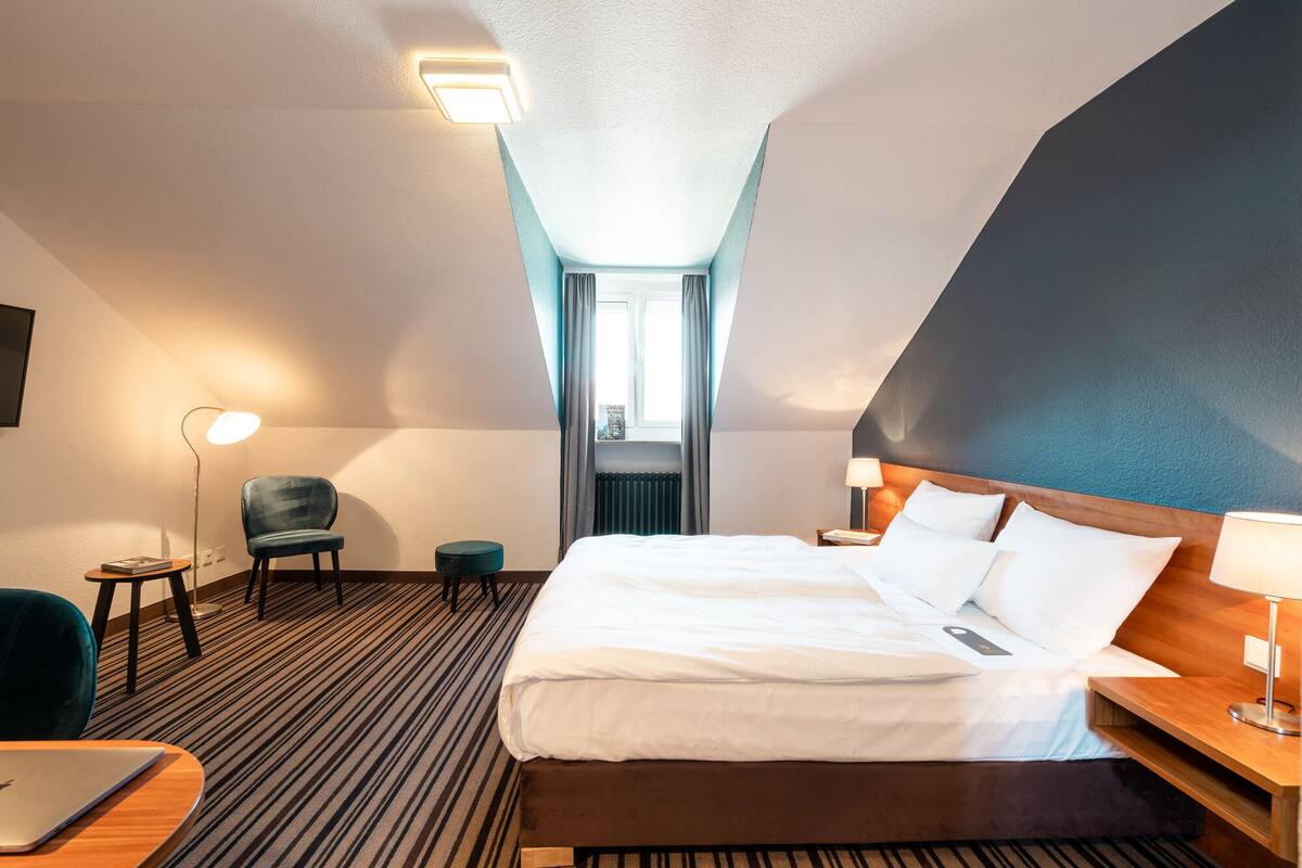A hotel room with a large bed, a desk with a laptop, a chair, a small table, and a nightstand with a lamp.