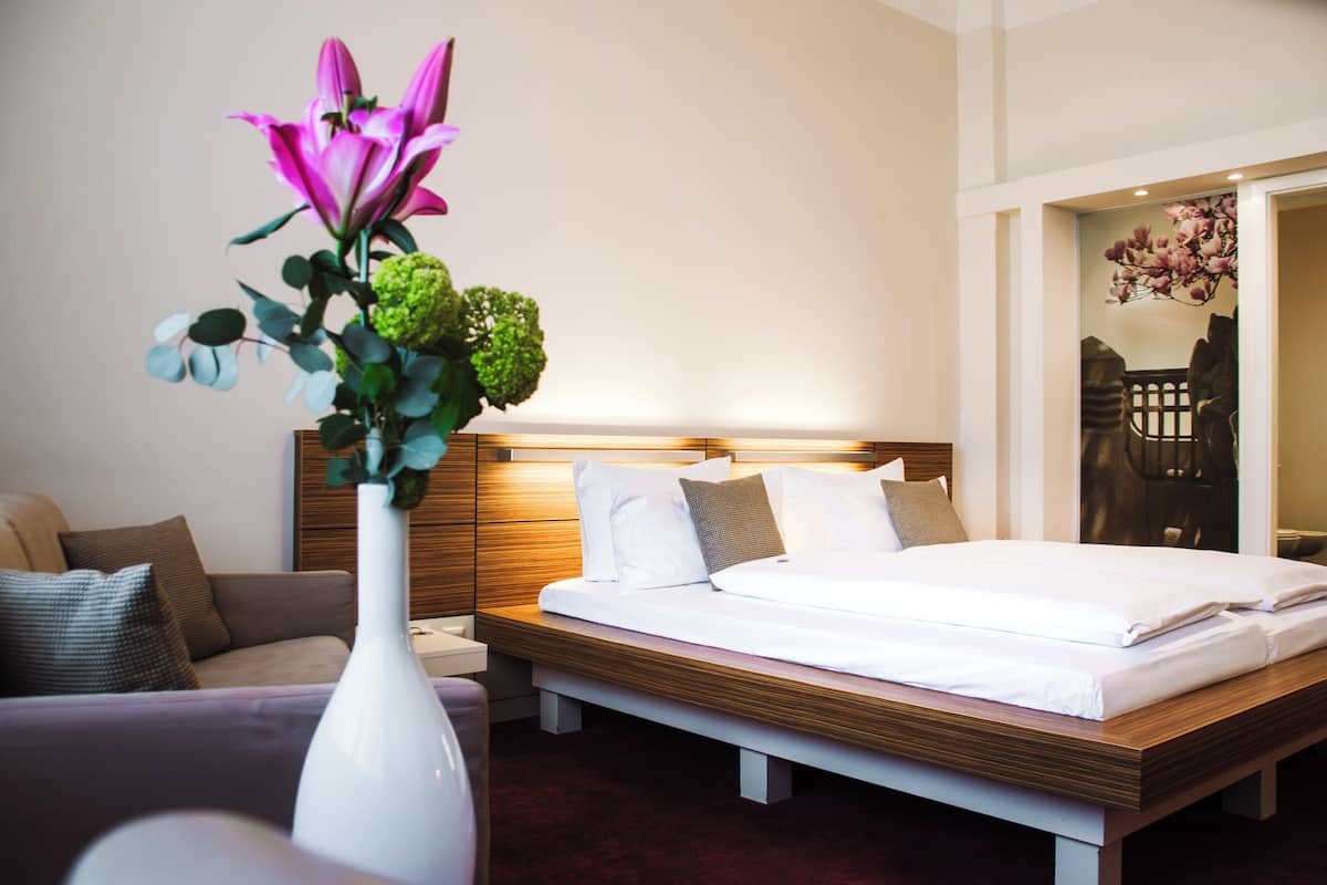 Quarto de hotel com uma cama, mesa de cabeceira, vaso branco com flores, sofá e cabeceira de madeira.