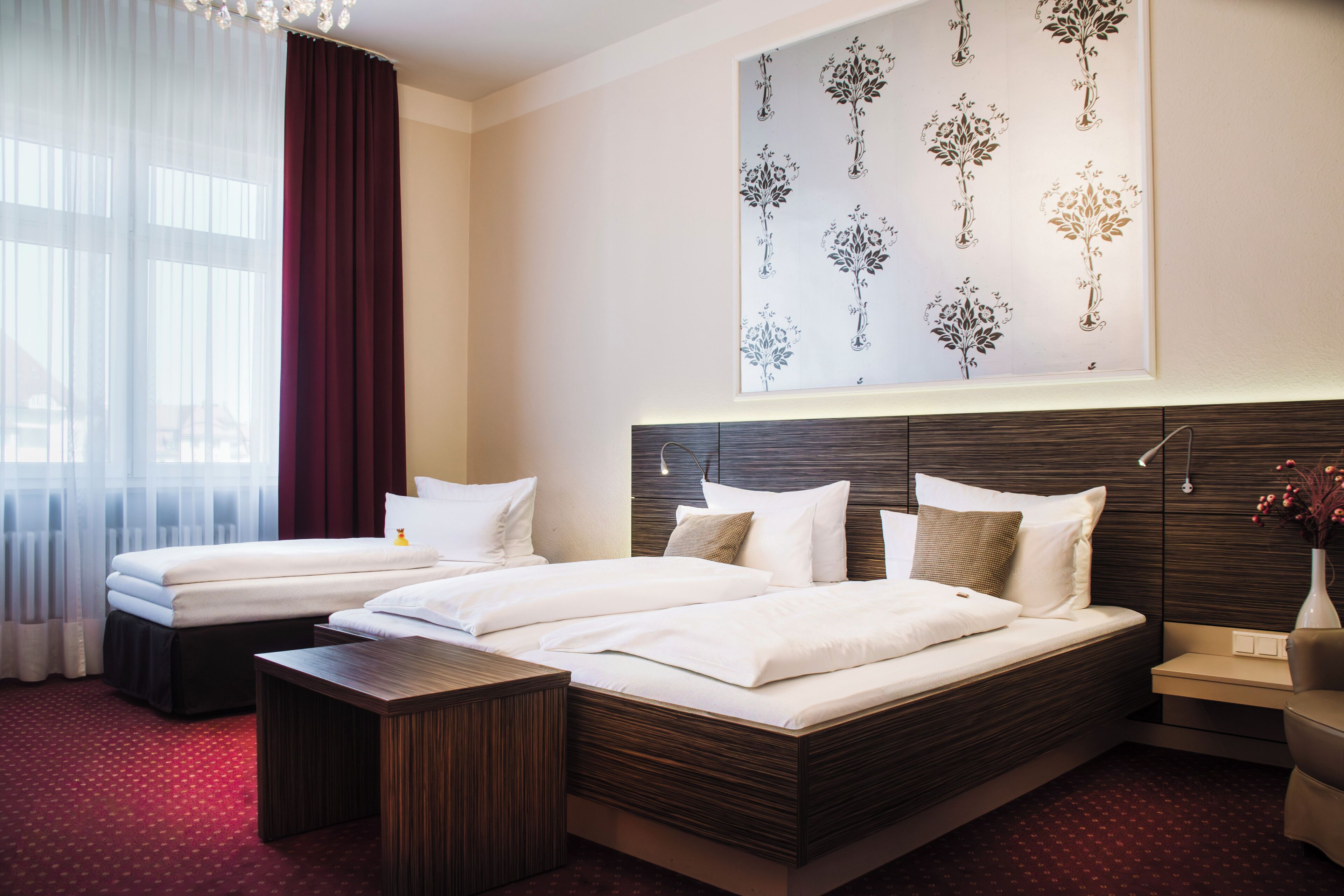 Habitación de hotel con dos camas, un ventanal con cortinas translúcidas, una obra de arte colgada en la pared y una mesita de noche con un florero.