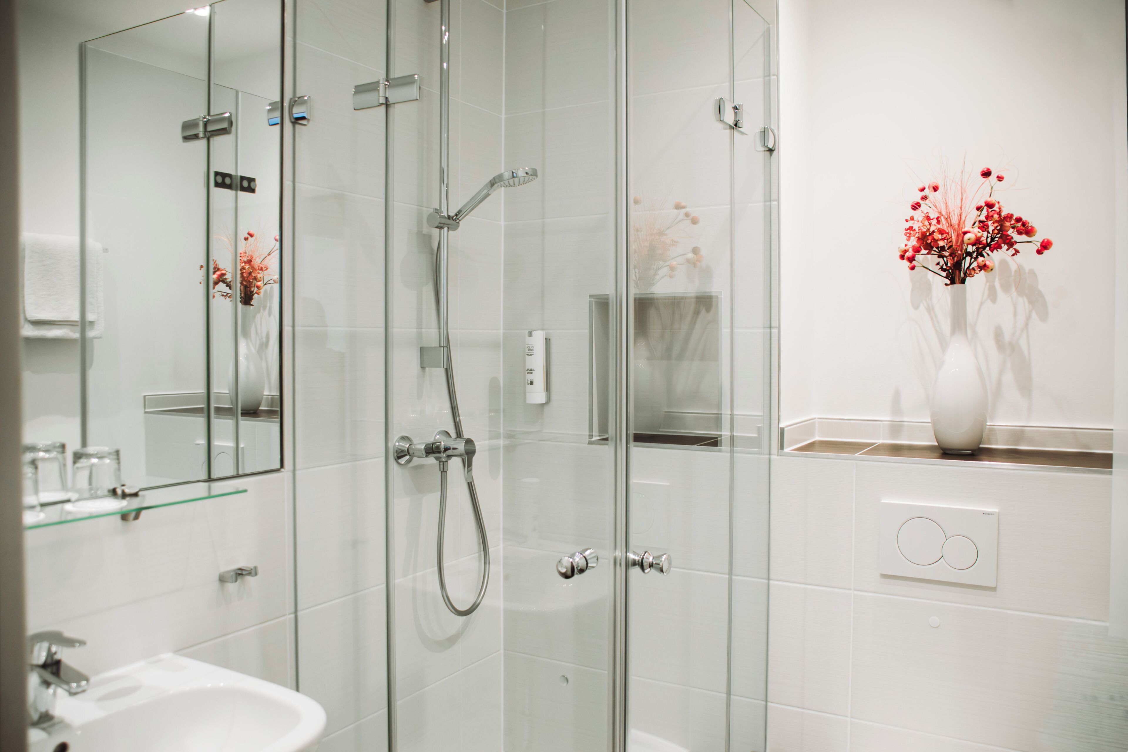 A modern bathroom with a glass-enclosed shower, a white sink, and a vase with red flowers on the wall.