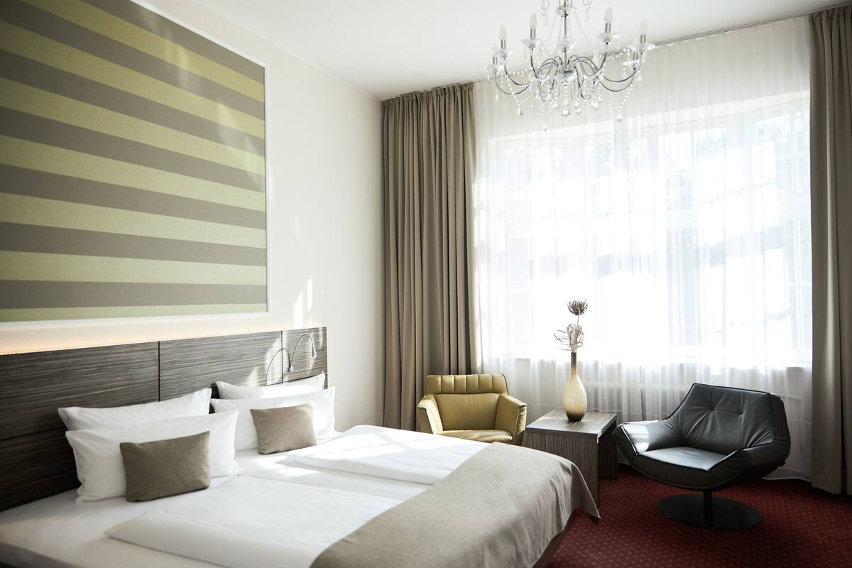 Quarto de hotel com uma cama grande, duas cadeiras, um candelabro e uma janela ampla com cortinas.