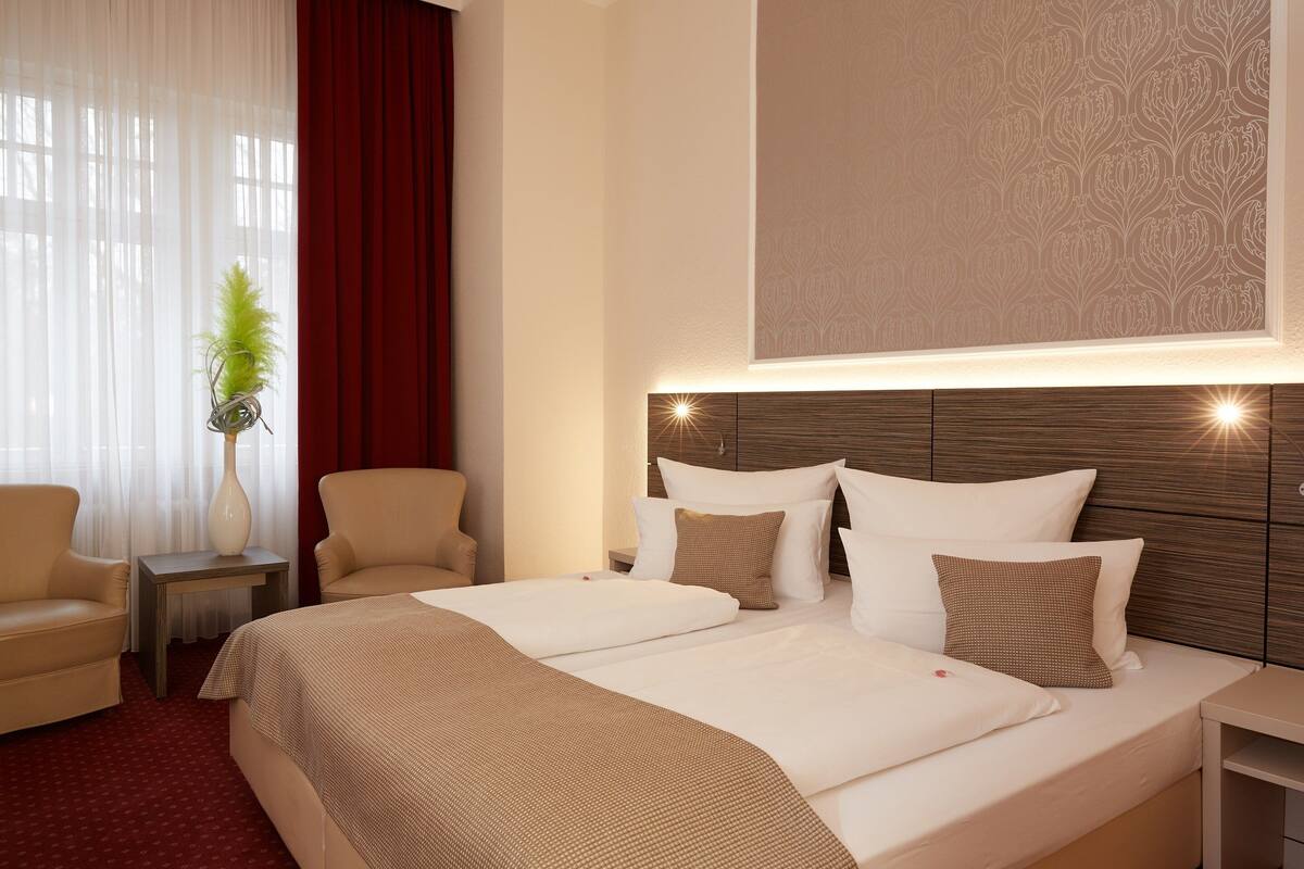 Quarto de hotel com uma cama grande, duas mesas de cabeceira, uma cadeira e um vaso de planta.