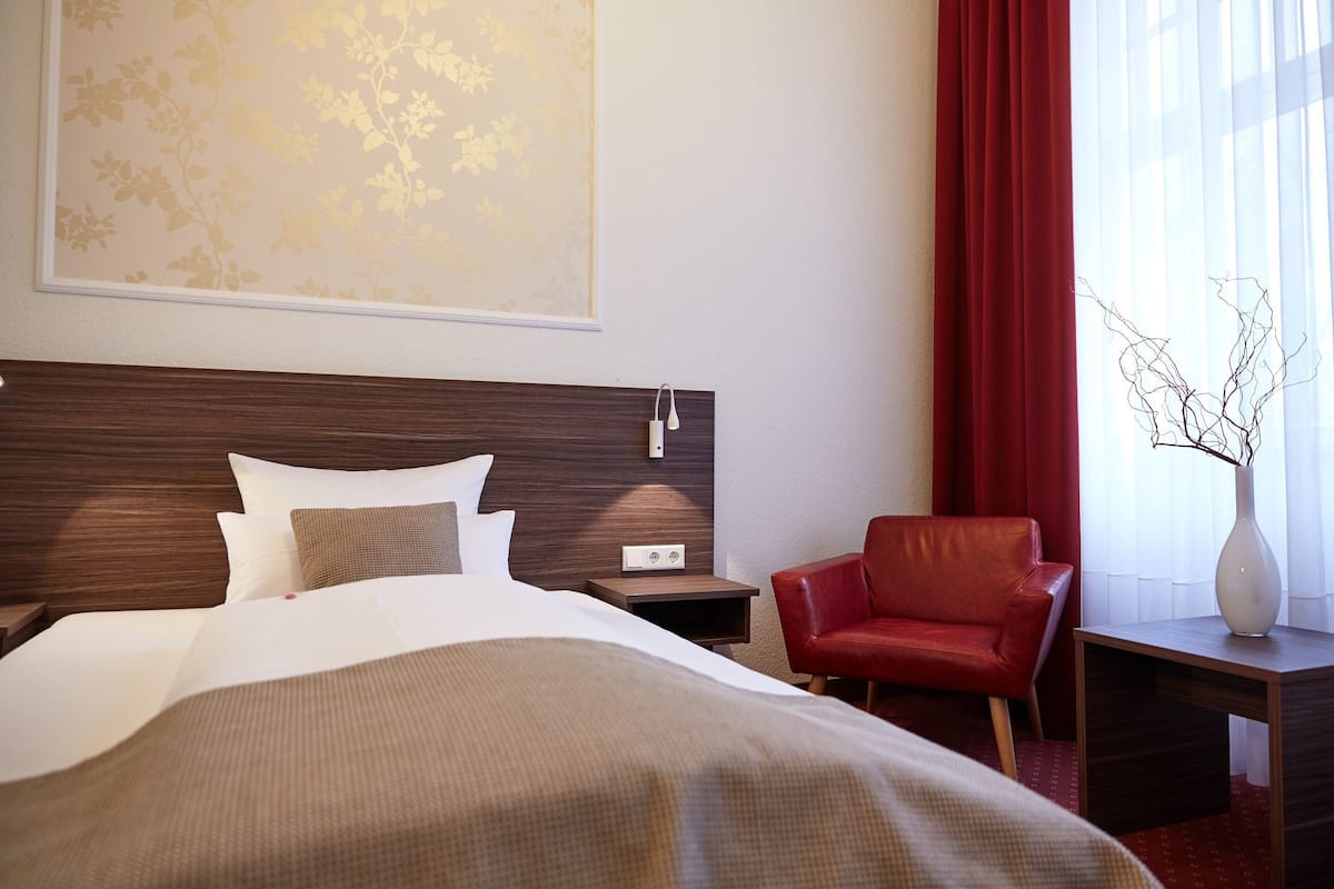 Quarto de hotel com uma cama grande, cabeceira de madeira, poltrona de couro vermelha, mesa de cabeceira com abajur e janela com cortinas vermelhas.